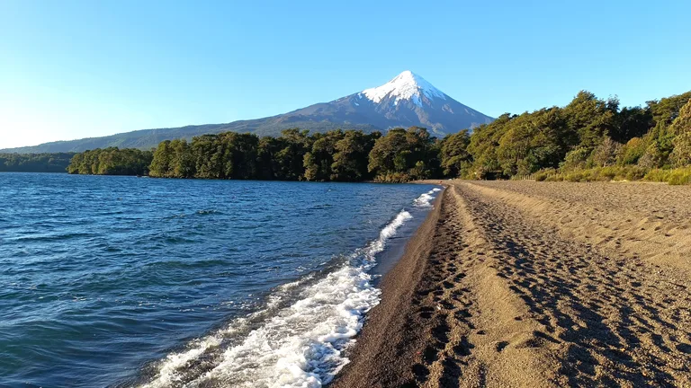 Playa de arena volcánica oscura en la orilla oriental del lago Llanquihue, con bosque nativo y el volcán Osorno nevado al fondo, en Ensenada
