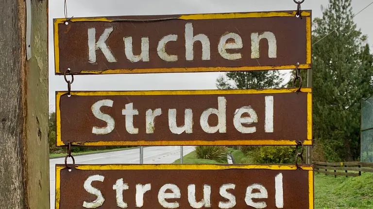 Letreros de madera y metal que ofrecen kuchen, strudel y streusel junto a un camino en Puerto Varas