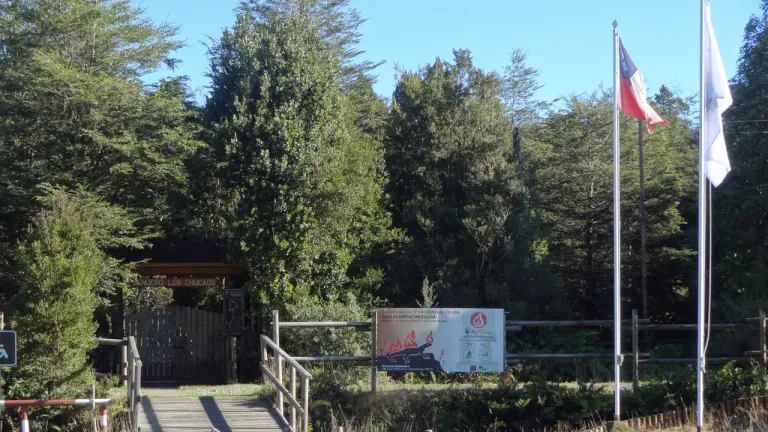 Portón de madera del sendero Los Chucaos, con bosque nativo y banderas, en la entrada del Monumento Natural Lahuen Ñadi