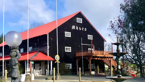 Fachada del Museo Antonio Felmer en Nueva Braunau: un granero de tejuelas oscuras con techo rojo a dos aguas y la palabra Museo en letras góticas, con el estanque y la fuente del jardín en primer plano