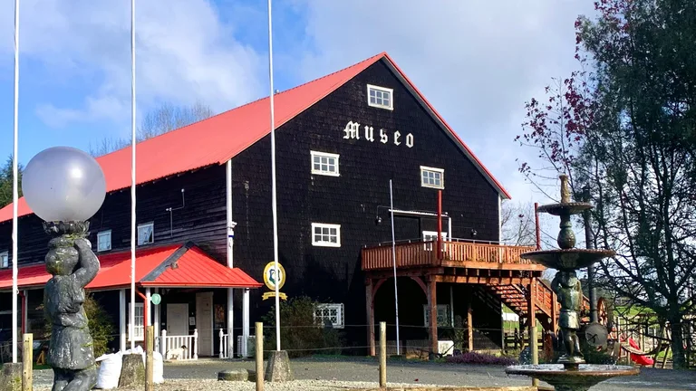 Fachada del Museo Antonio Felmer en Nueva Braunau: un granero de tejuelas oscuras con techo rojo a dos aguas y la palabra Museo en letras góticas, con el estanque y la fuente del jardín en primer plano
