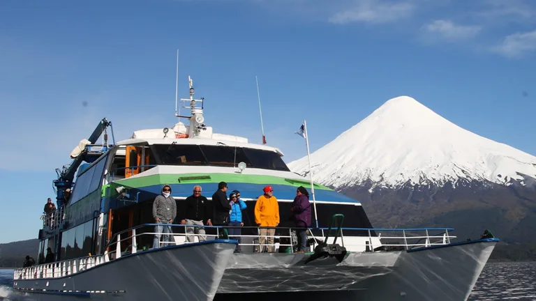 Catamarán con pasajeros en cubierta navegando el lago Todos los Santos hacia Peulla, con el volcán Osorno nevado al fondo